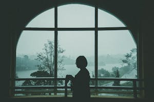 A silhouette of a woman standing by a large arched window with a scenic view outdoors. Moody and artistic.